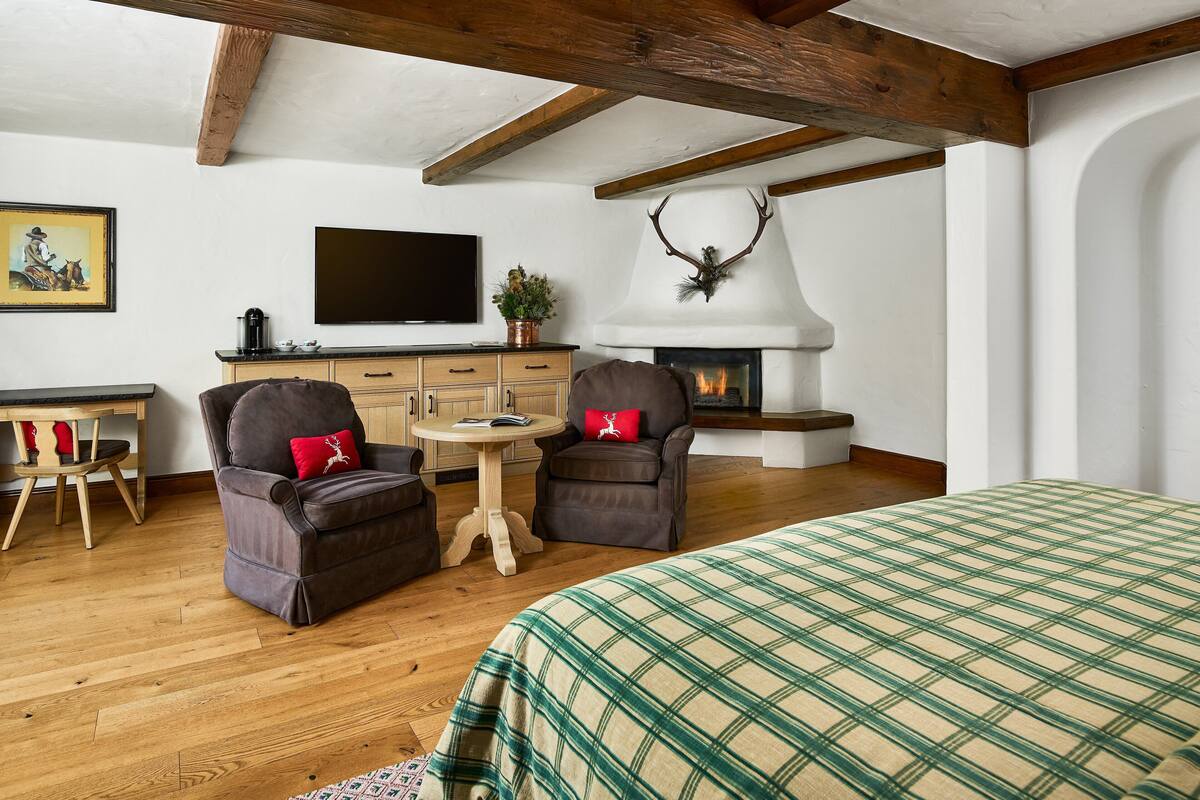 A cozy room with a fireplace, two armchairs, a bed with a plaid bedspread, a television, and a painting on the wall.