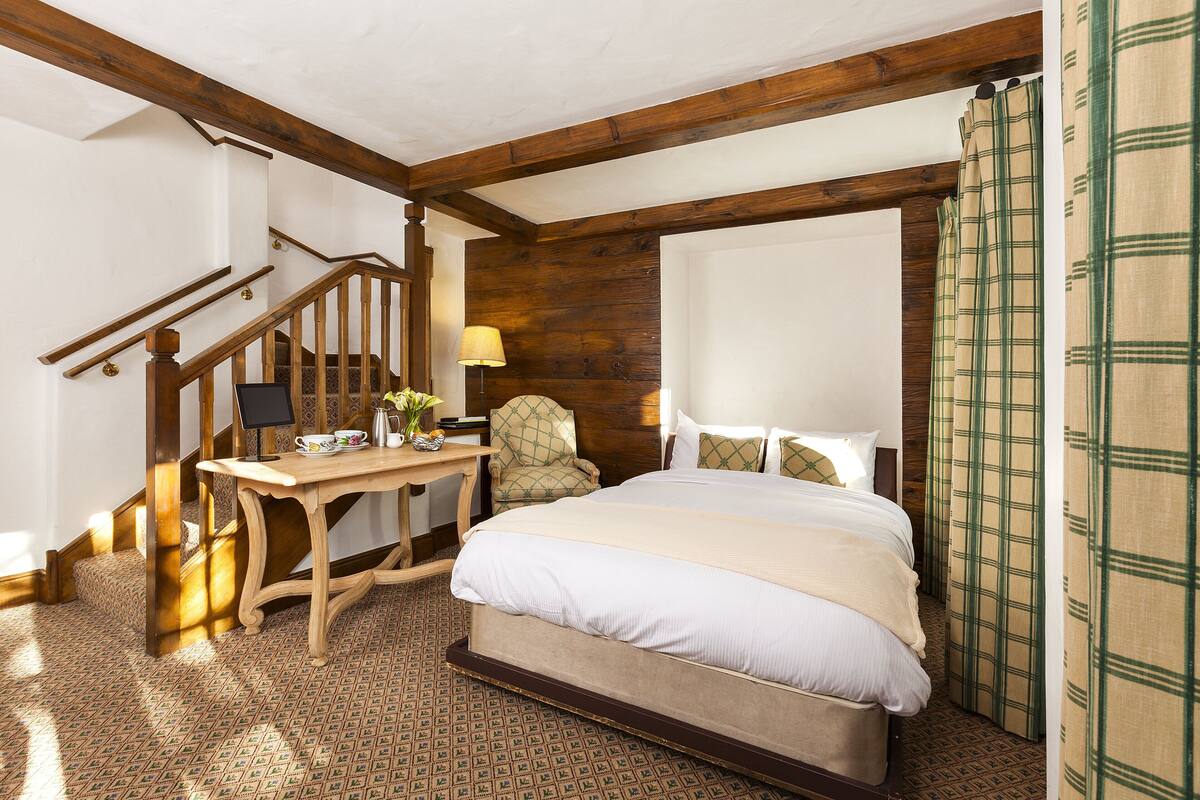 A bedroom with a wooden staircase, a bed with white bedding, a wooden desk with a lamp, and a patterned carpet.