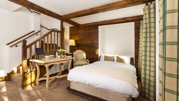 A bedroom with a wooden staircase, a bed with white bedding, a wooden desk with a lamp, and a patterned carpet.