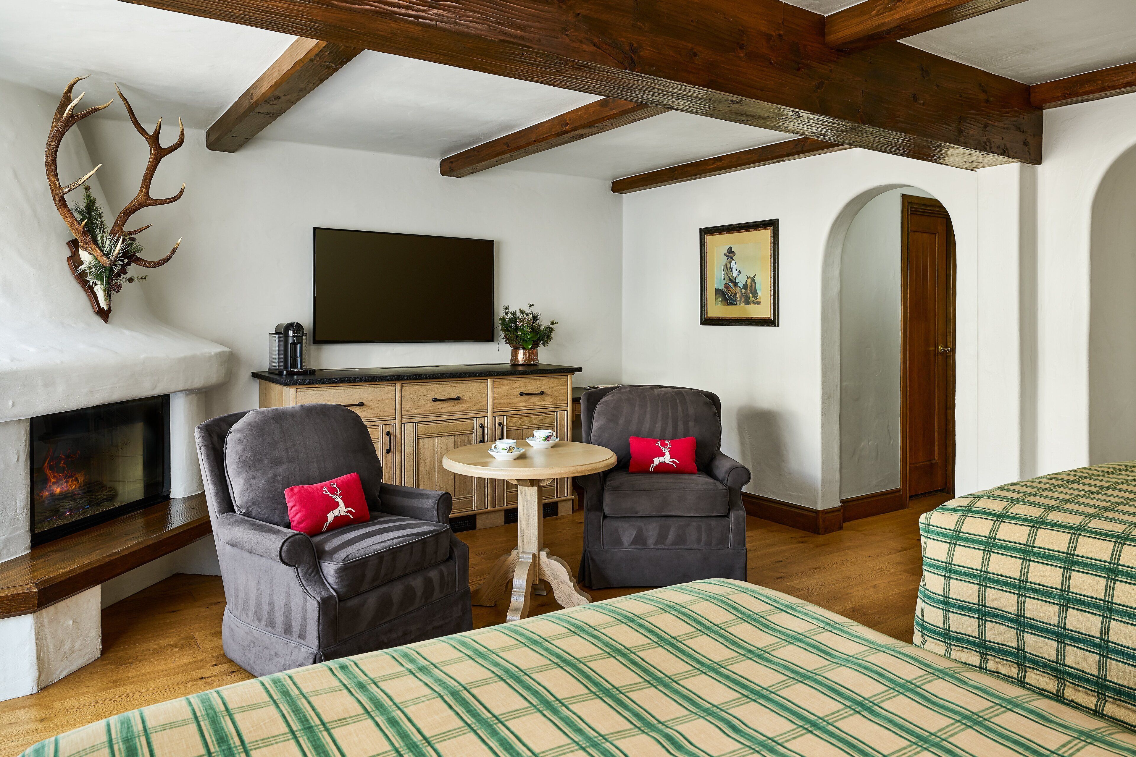 Ein gemütliches Zimmer mit Kamin, zwei Sesseln, einem kleinen Tisch und einem Bett mit karierter Bettwäsche.