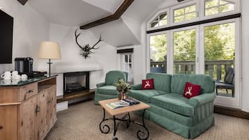 Un salon chaleureux avec une cheminée, un canapé vert, un meuble en bois et une grande fenêtre donnant sur la forêt.