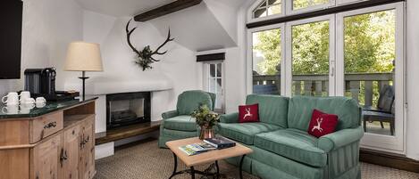 A cozy living room with a fireplace, green sofa, wooden cabinet, and a large window with a view of trees.