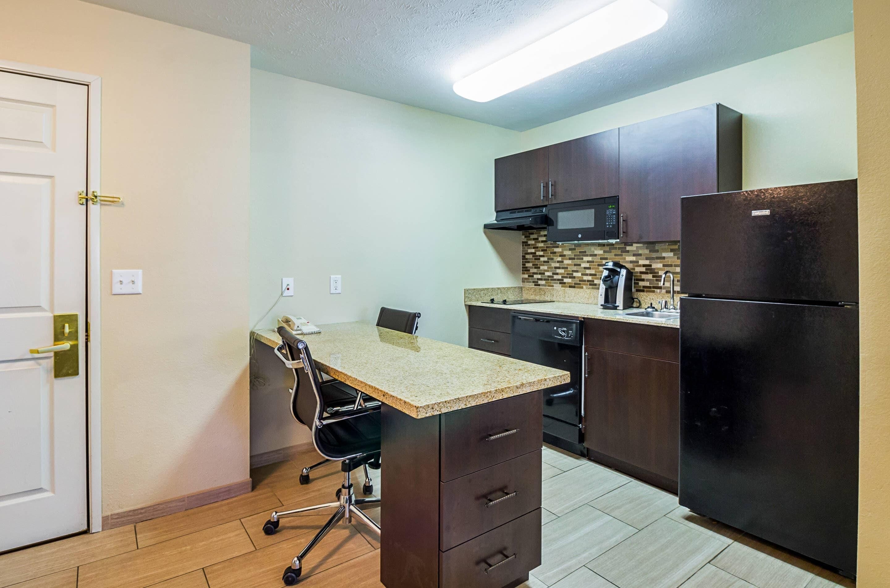 A compact kitchen with dark cabinets, a microwave, a refrigerator, a small countertop, and a chair.