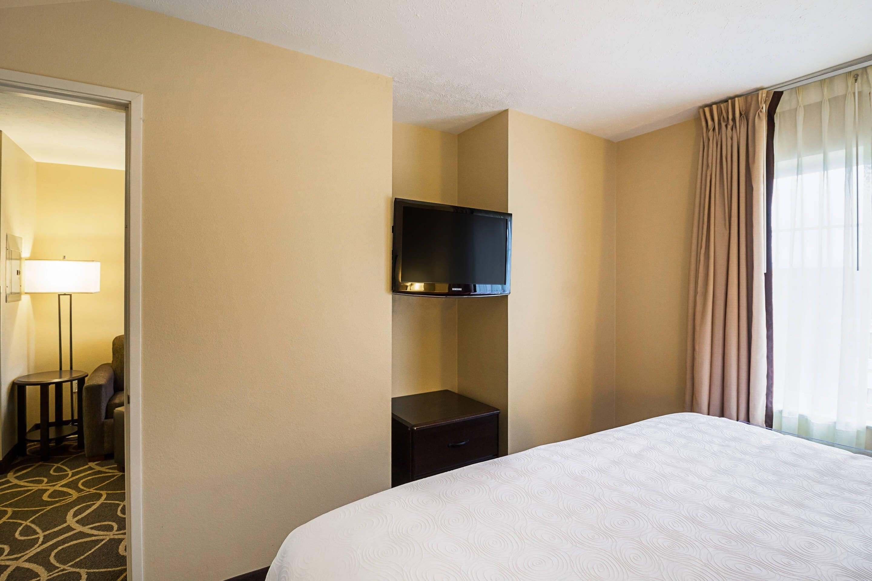 A hotel room with a bed, a television mounted on the wall, a small table with a chair, and a lamp.