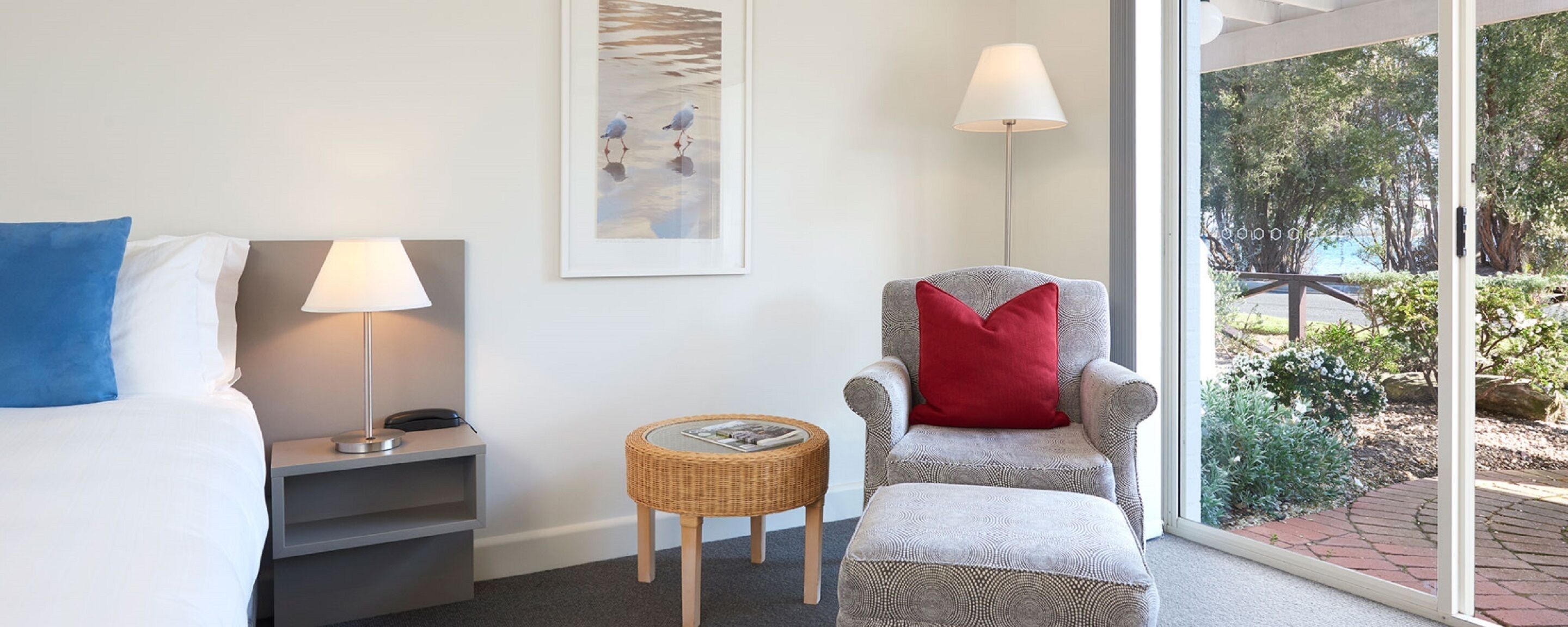 A hotel room with a bed, a chair with a red pillow, a lamp, a small wicker table, and a framed picture on the wall.