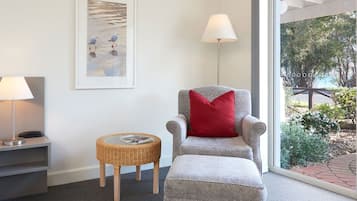 A hotel room with a bed, a chair with a red pillow, a lamp, a small wicker table, and a framed picture on the wall.