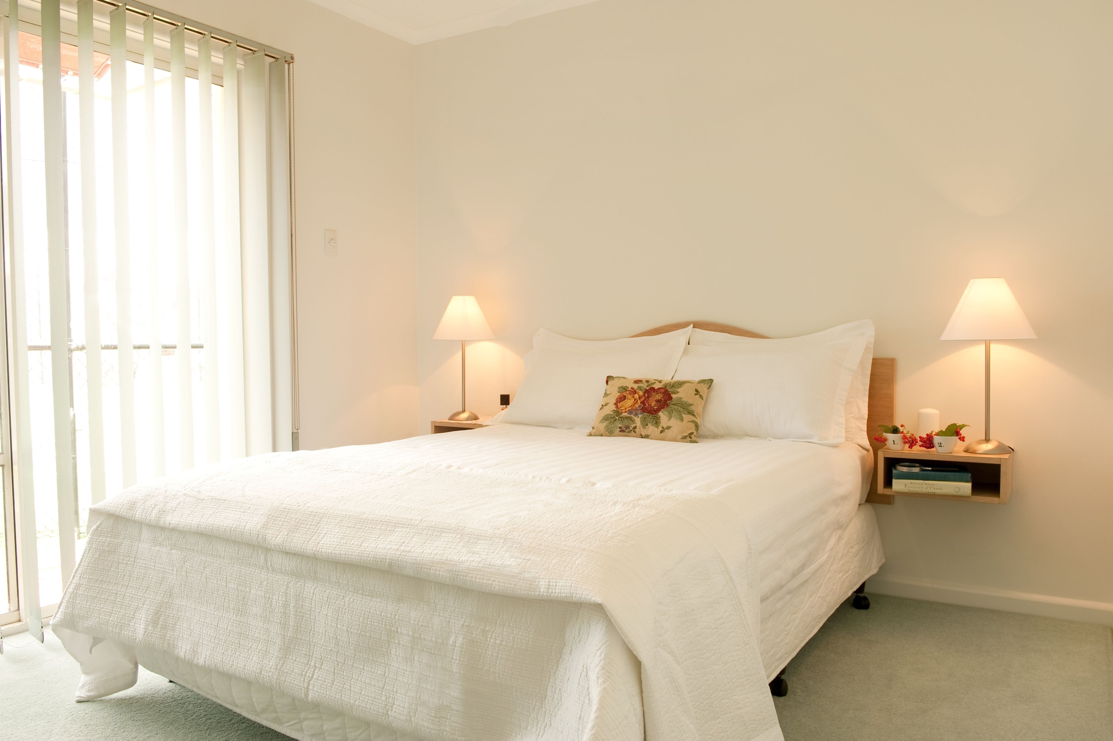 A neatly made bed with white bedding and a floral pillow, flanked by bedside tables with lamps and a small shelf with decorative items.