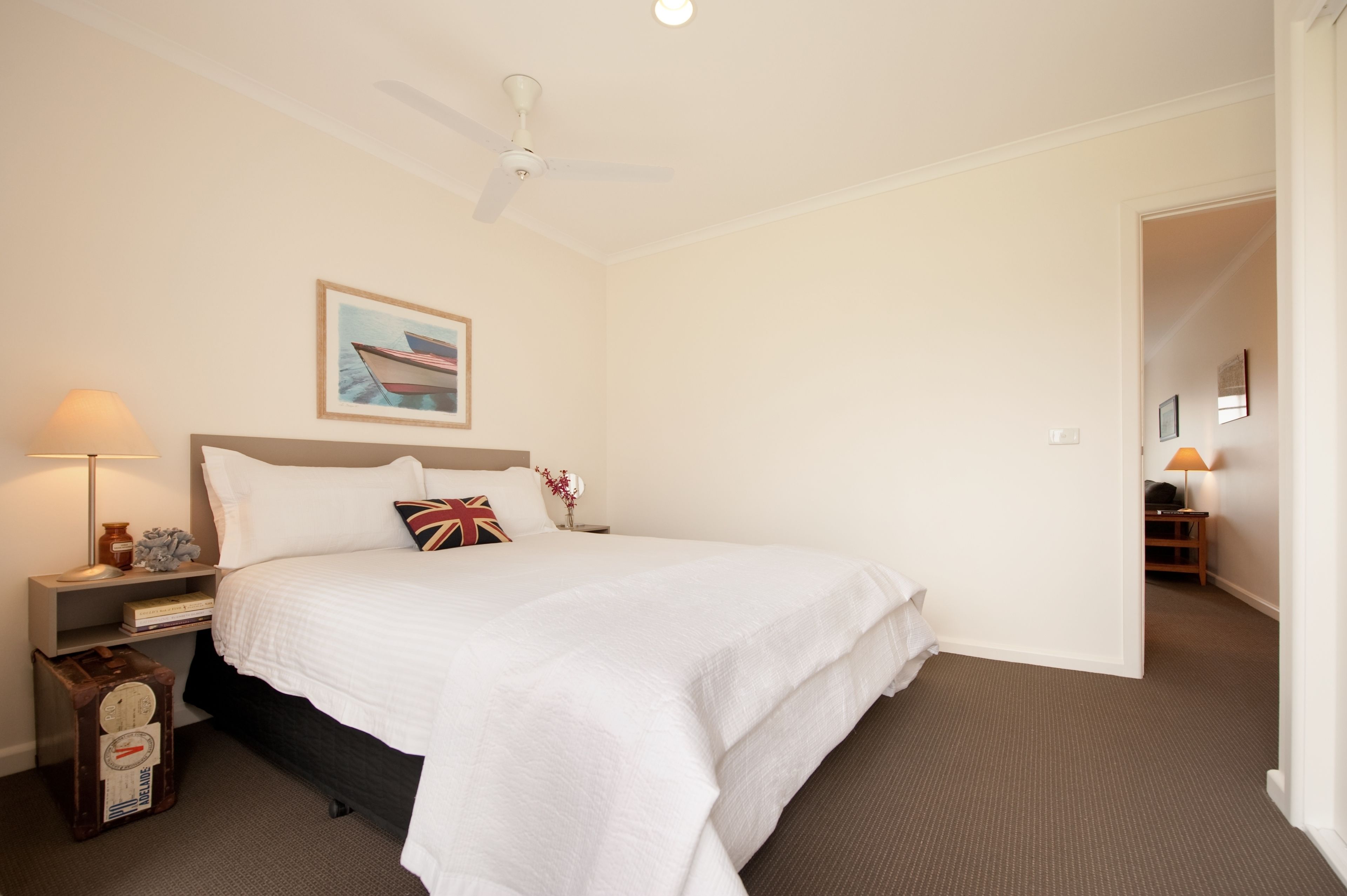 Una habitación de hotel con una cama, una mesita de noche, una lámpara, una maleta, un ventilador de techo y un cuadro en la pared.