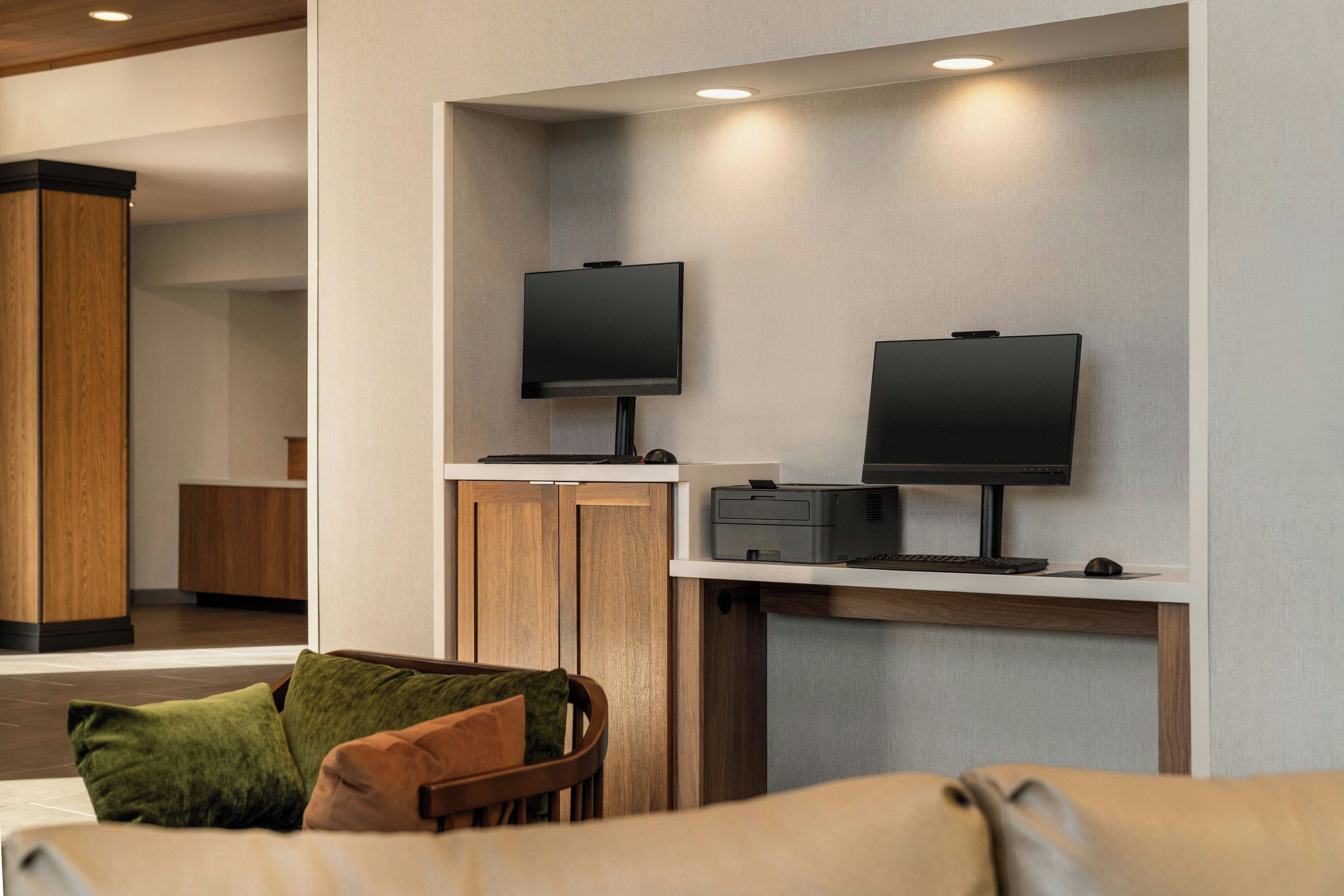 Un hall d’hôtel moderne doté d’un espace salon, de deux téléviseurs à écran plat fixés au mur, d’une imprimante et d’un meuble en bois.