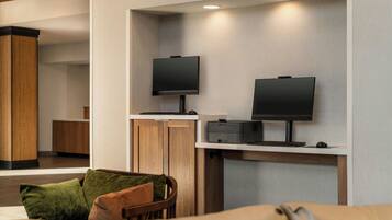 Un hall d’hôtel moderne doté d’un espace salon, de deux téléviseurs à écran plat fixés au mur, d’une imprimante et d’un meuble en bois.