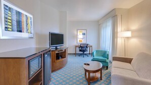 A hotel room with a TV, desk, chair, sofa, and a patterned floor. - Fairfield Inn & Suites by Marriott San Francisco Airport (Millbrae)