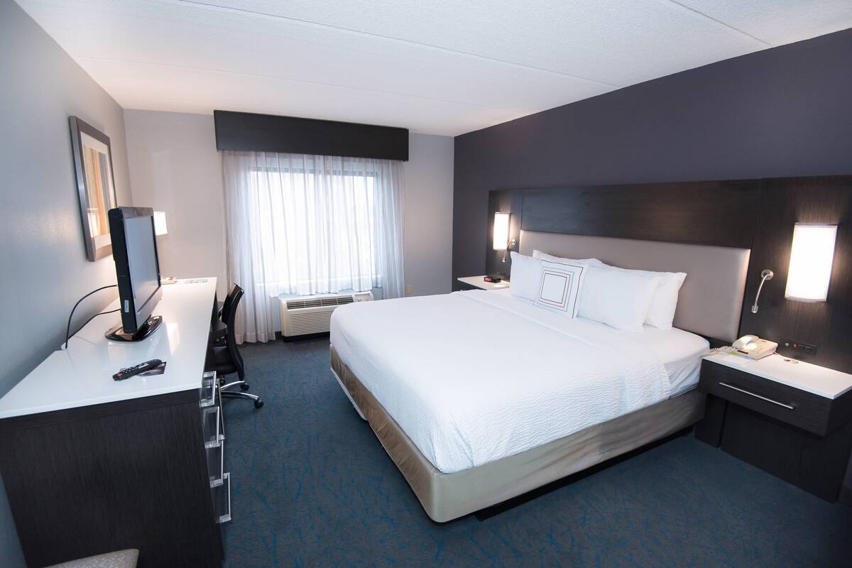Een hotelkamer met een groot bed, een bureau met een computer, een stoel, een nachtkastje met een lamp en een raam met gordijnen.