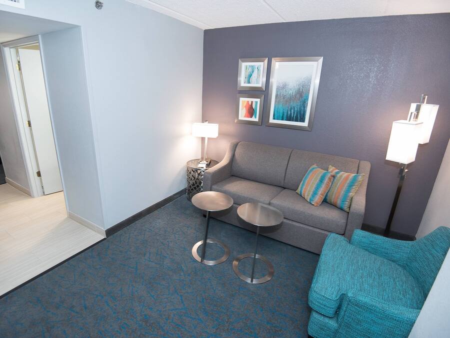 A hotel room with a grey sofa, two small round tables, a teal armchair, and a bed in the adjacent room.