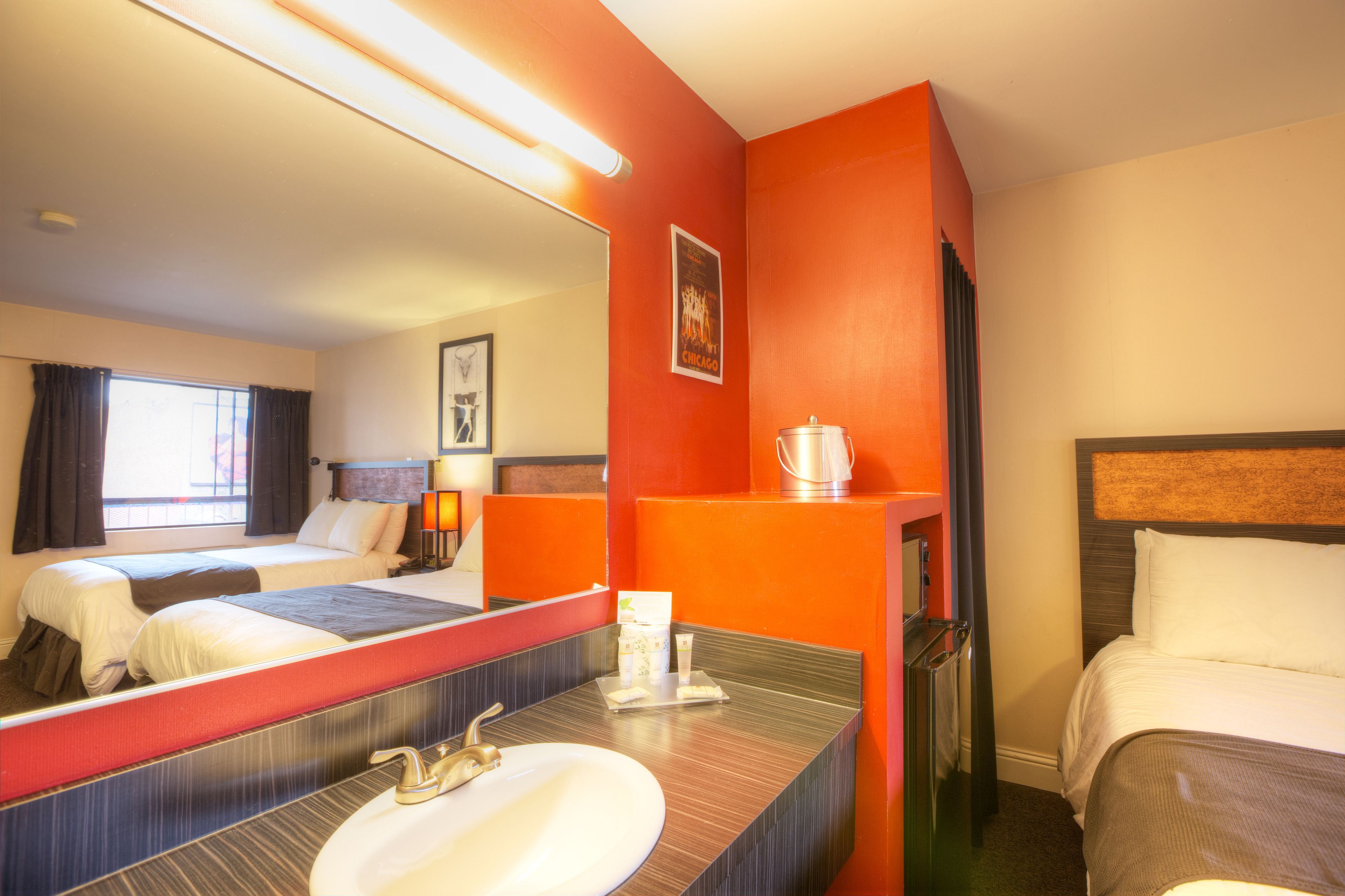Una camera d'albergo con due letti, un grande specchio, un bagno con lavandino e una parete rossa a contrasto.