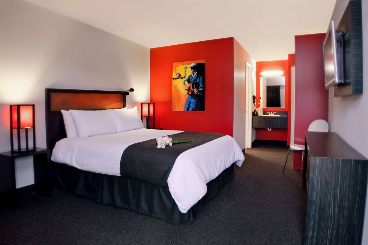 A hotel room with a large bed, a red accent wall, a painting, a desk, and a TV.