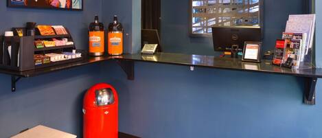 A reception area with a desk, two coffee dispensers, a trash bin, a small table, and a painting on the wall.