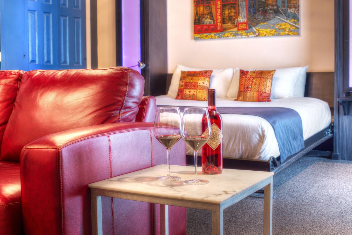 A hotel room with a red leather sofa, a bed with patterned pillows, and two glasses of red wine on a small table.