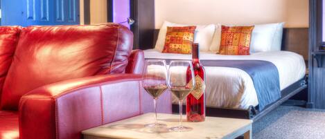 A hotel room with a red leather sofa, a bed with patterned pillows, and two glasses of red wine on a small table.