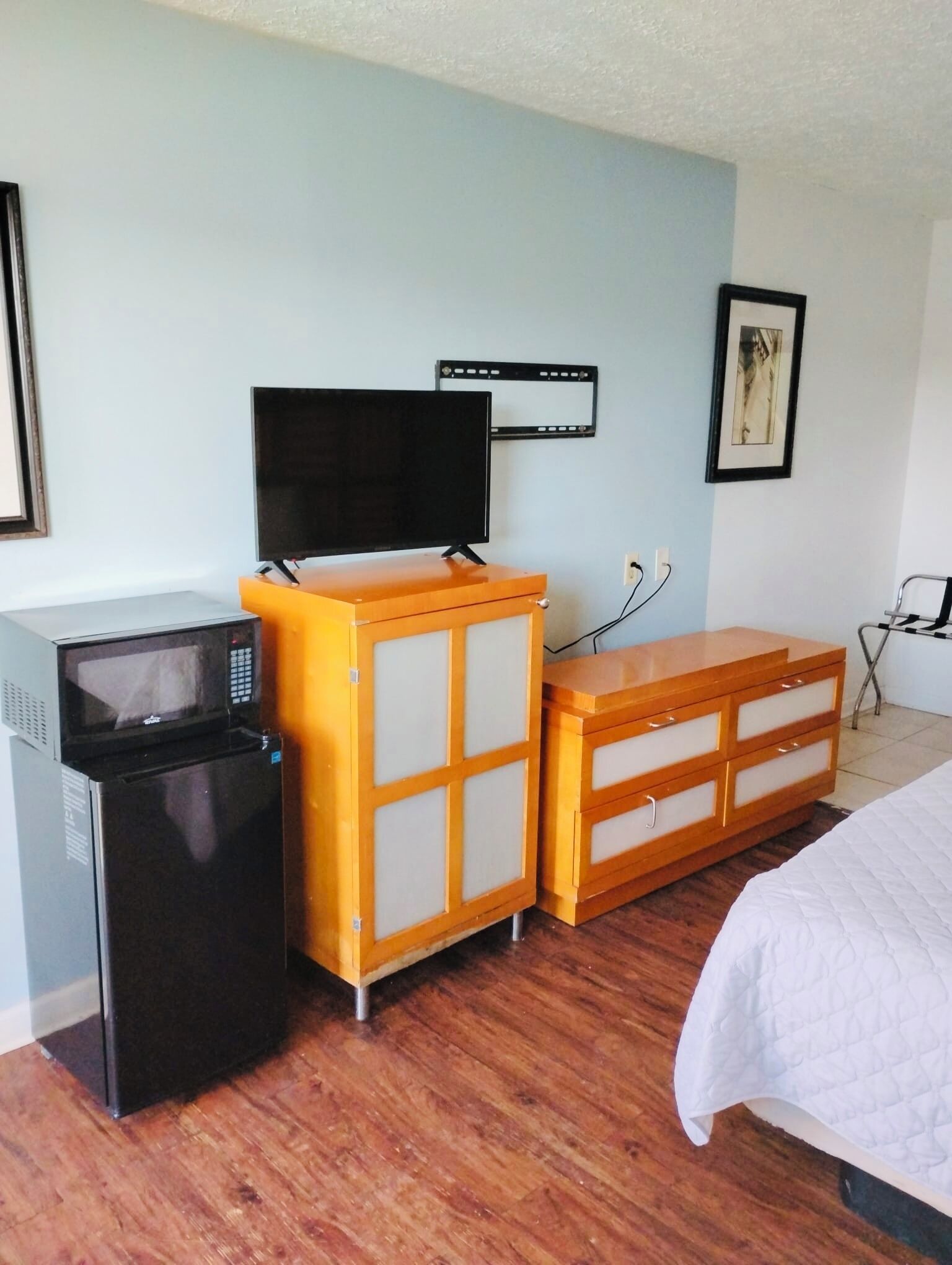 A hotel room with a bed, a wooden dresser, a microwave, a television, and a small refrigerator.