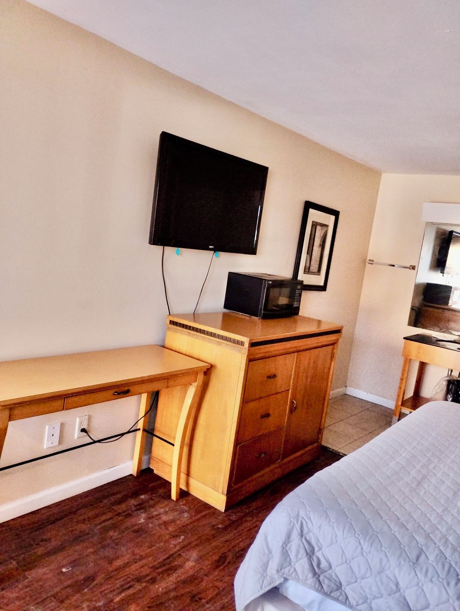 A hotel room with a wooden desk, a television mounted on the wall, a bed with a quilted cover, and a refrigerator.