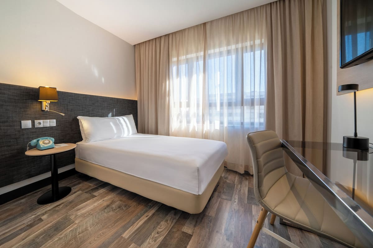 A hotel room with a bed, a desk with a lamp, a chair, a TV, and a window with curtains.