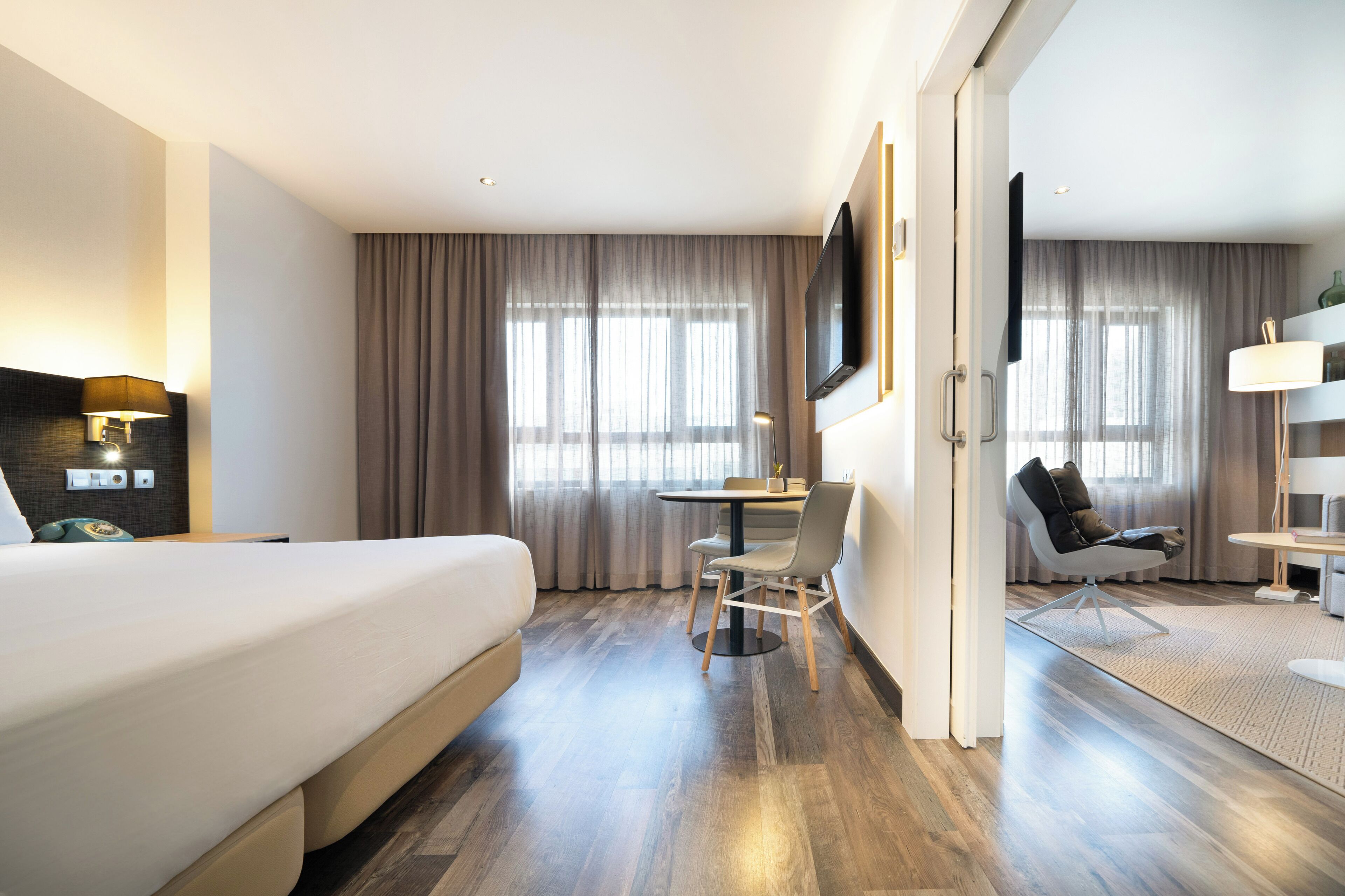Et hotelværelse med seng, skrivebord, stol, fjernsyn og store vinduer med gardiner.