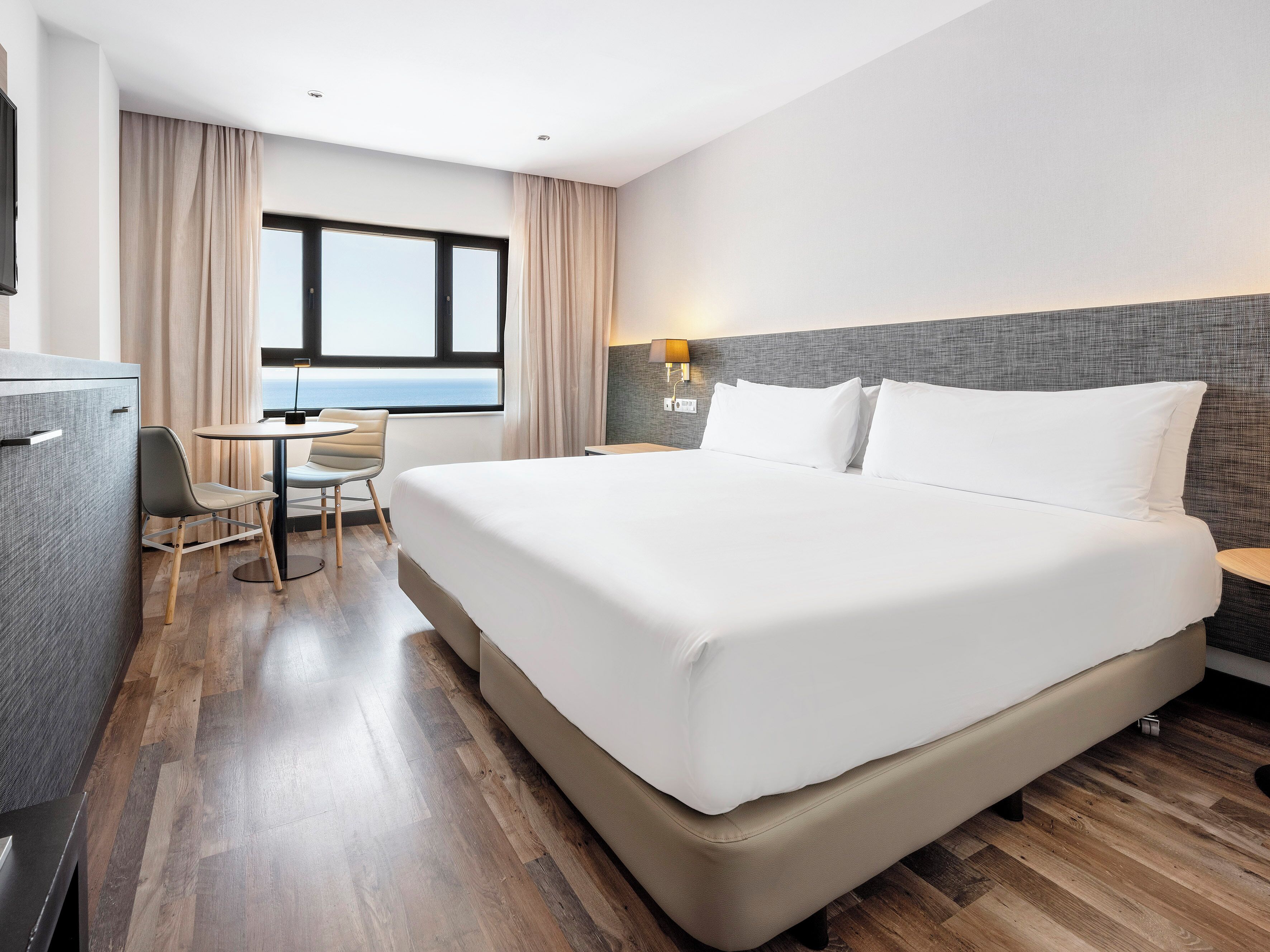 A hotel room with a large bed, a desk with a chair, a small table, and a view of the ocean through a window.