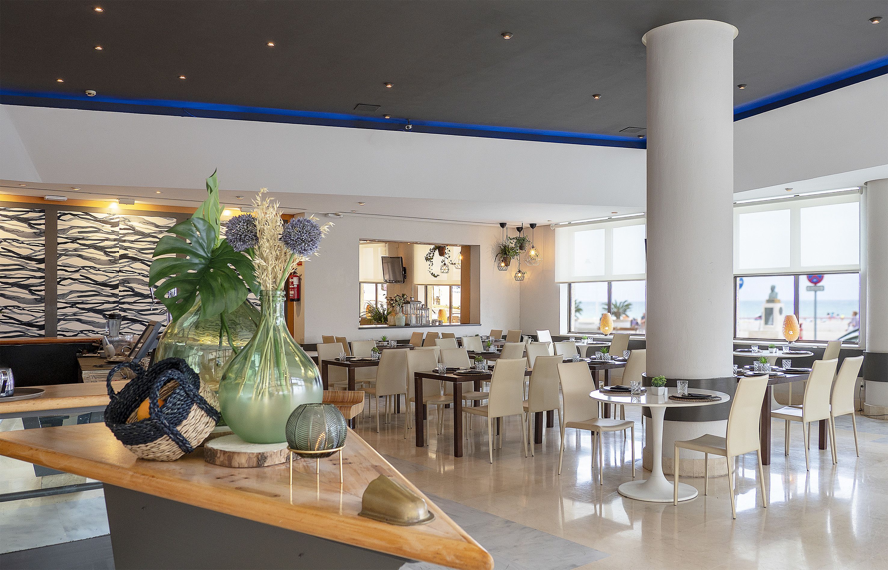 Un amplio vestíbulo de hotel con diseño moderno, que incluye un mostrador de recepción grande, varias mesas de comedor y vistas a la playa a través de amplios ventanales.