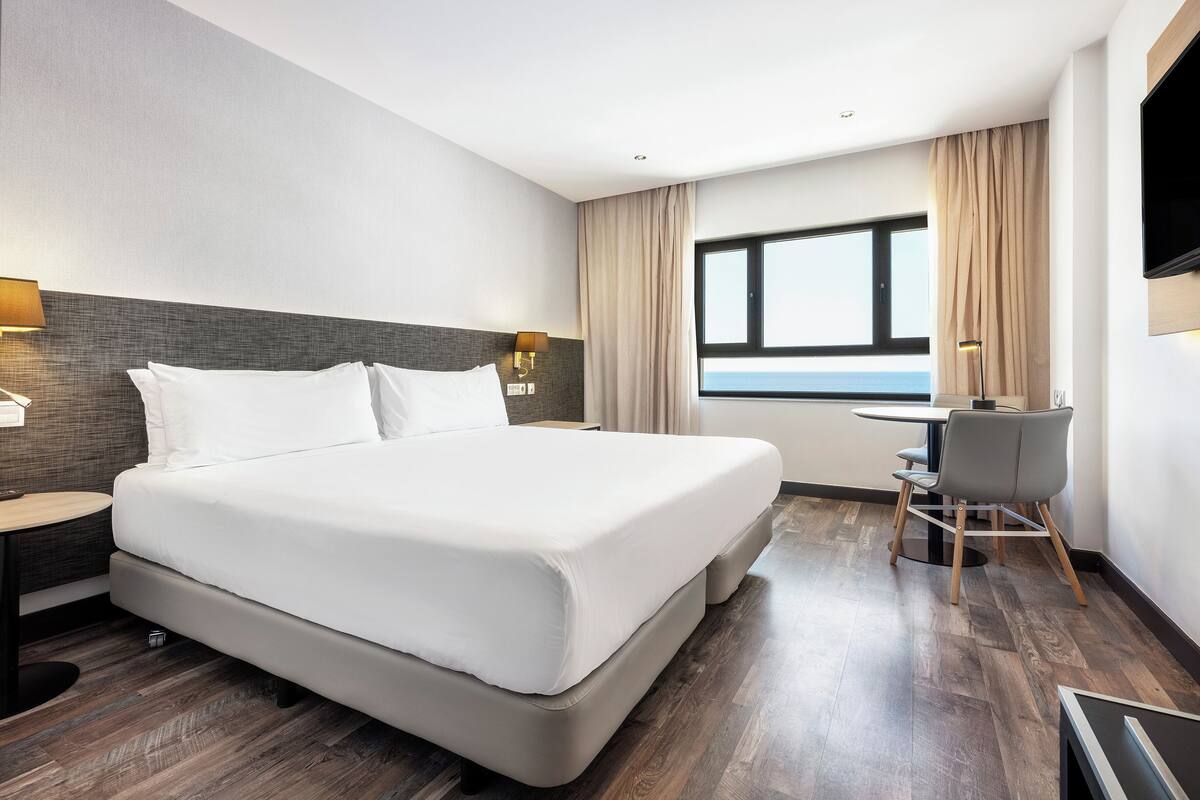 A hotel room with a large bed, a desk with a chair, a window with a view, and a TV mounted on the wall.