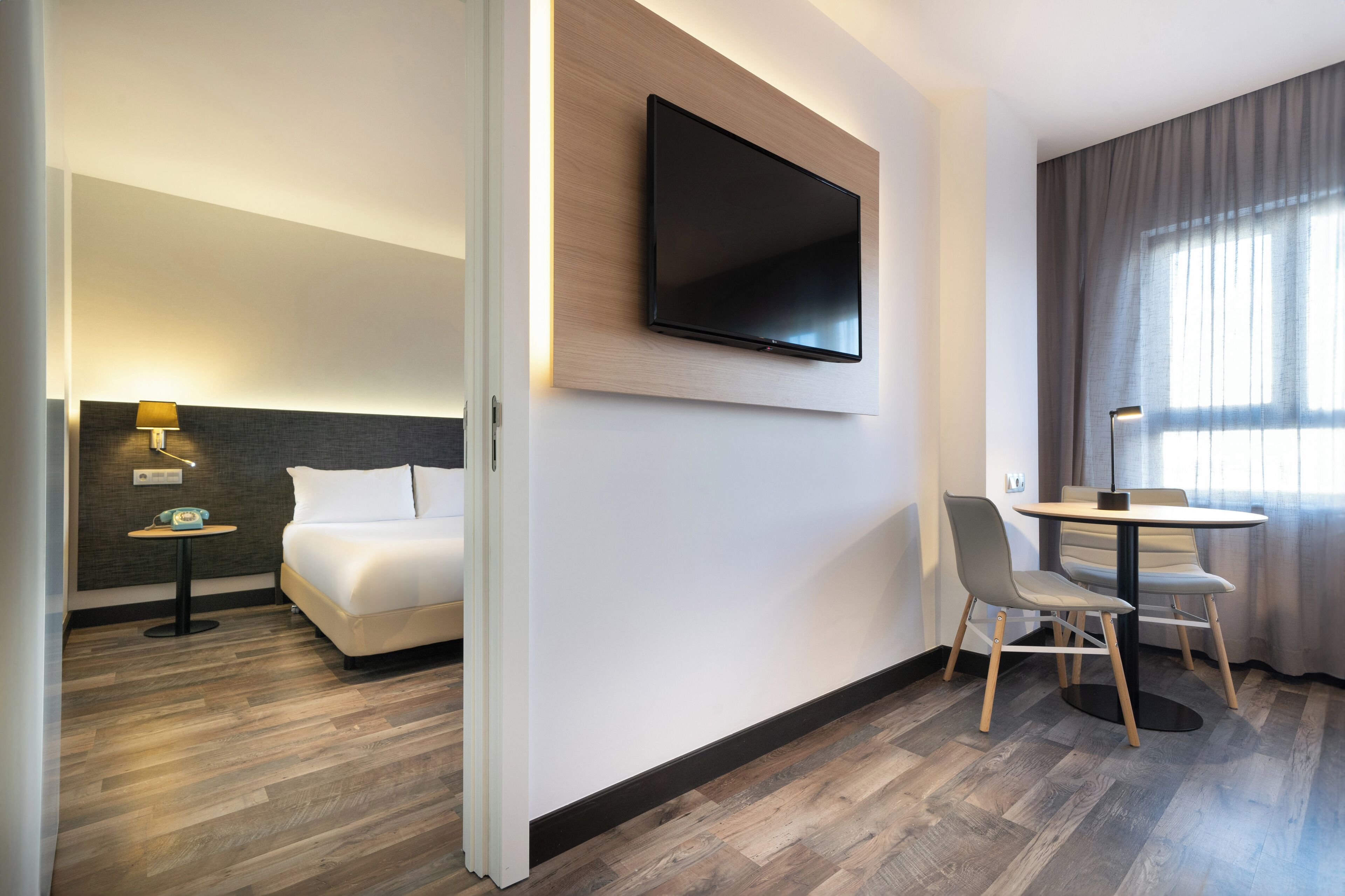 Ein Hotelzimmer mit einem Bett, einem an der Wand montierten Fernseher, einem kleinen Tisch mit Lampe sowie einem Essbereich mit Stühlen und Tisch.