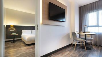 Habitación de hotel con cama, televisión montada en la pared, una mesita de noche con lámpara y un rincón comedor con sillas y mesa.