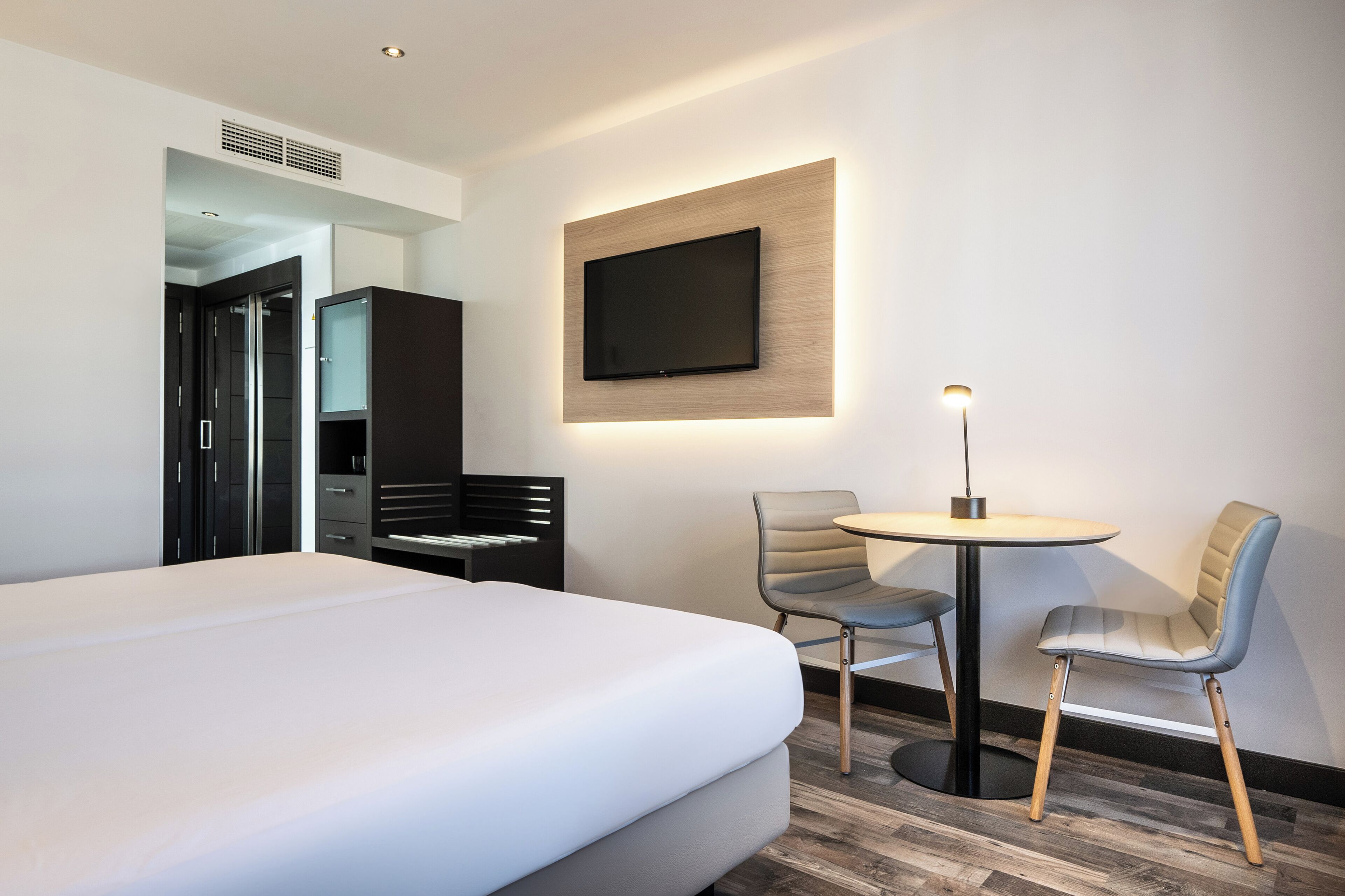 A modern hotel room with a large bed, a TV mounted on the wall, a small round table, and two chairs.