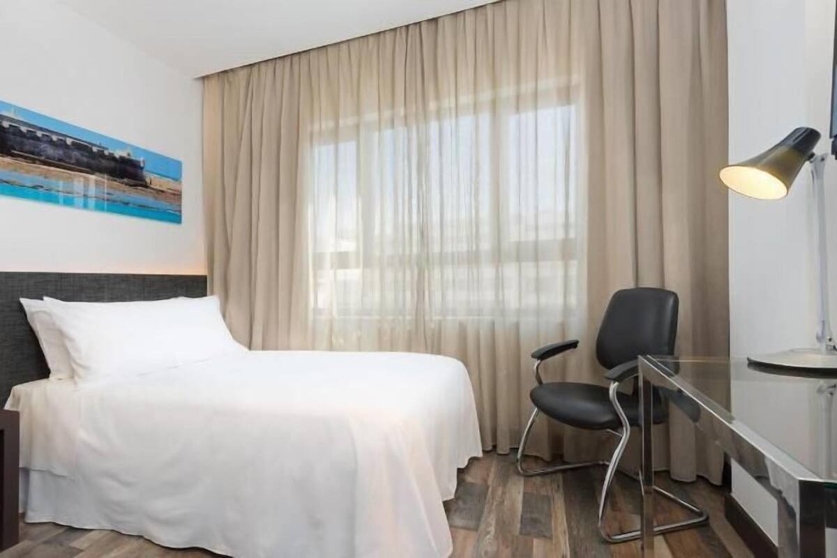 A hotel room with a bed, a desk, a chair, a lamp, a painting on the wall, and a window with curtains.