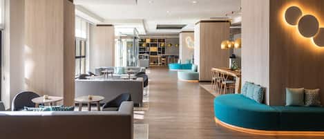 A modern hotel lobby with seating areas, a reception desk, and a visible kitchen in the background.