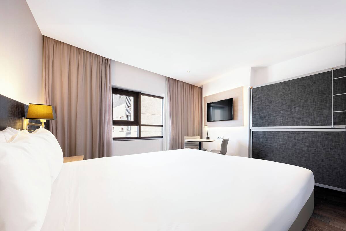 A hotel room with a large bed, a desk with a chair, a TV mounted on the wall, and a window with curtains.