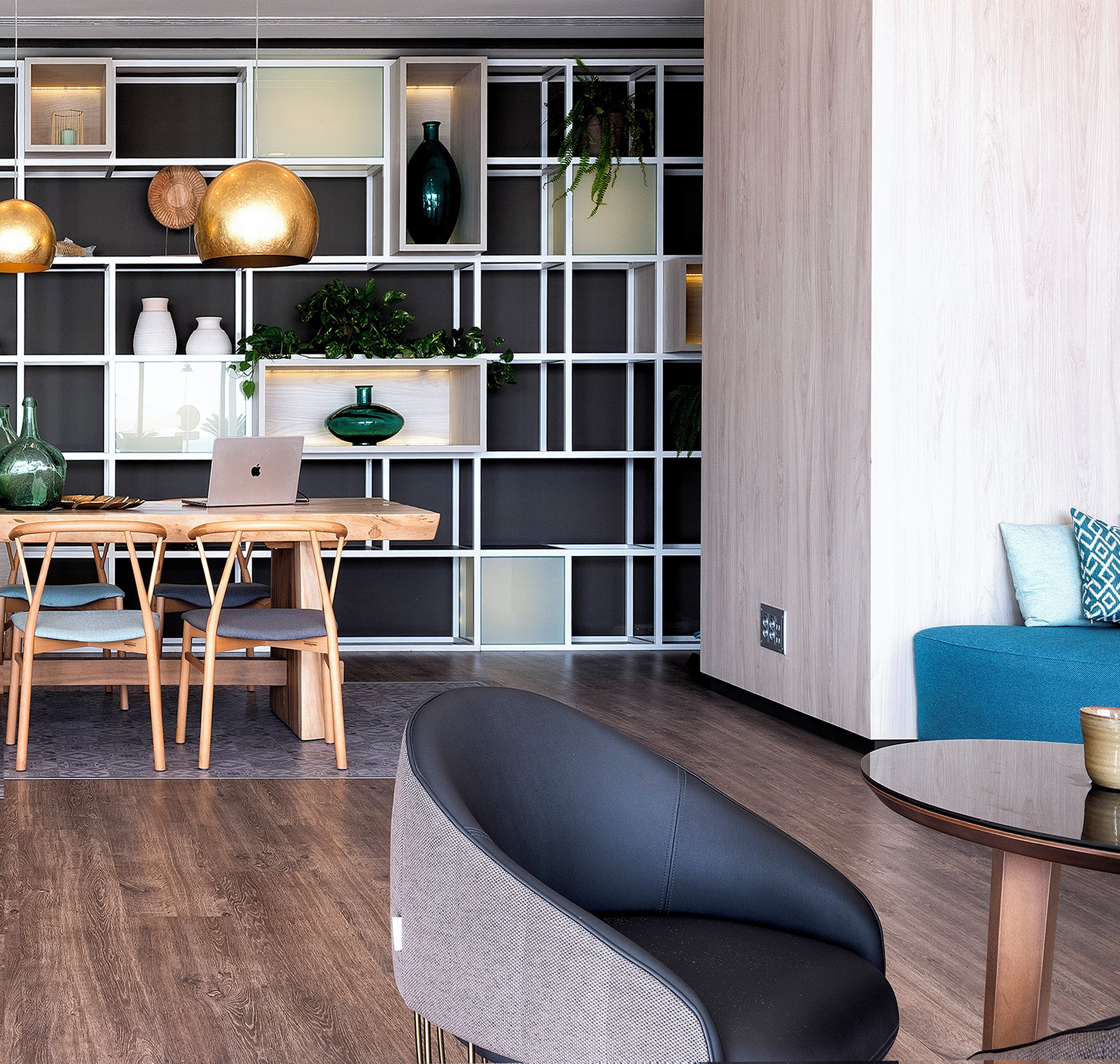 Una sala moderna con una mesa de comedor de madera, dos sillas, un sofá negro y una mesa redonda de centro. Se observa una estantería con objetos decorativos y plantas.