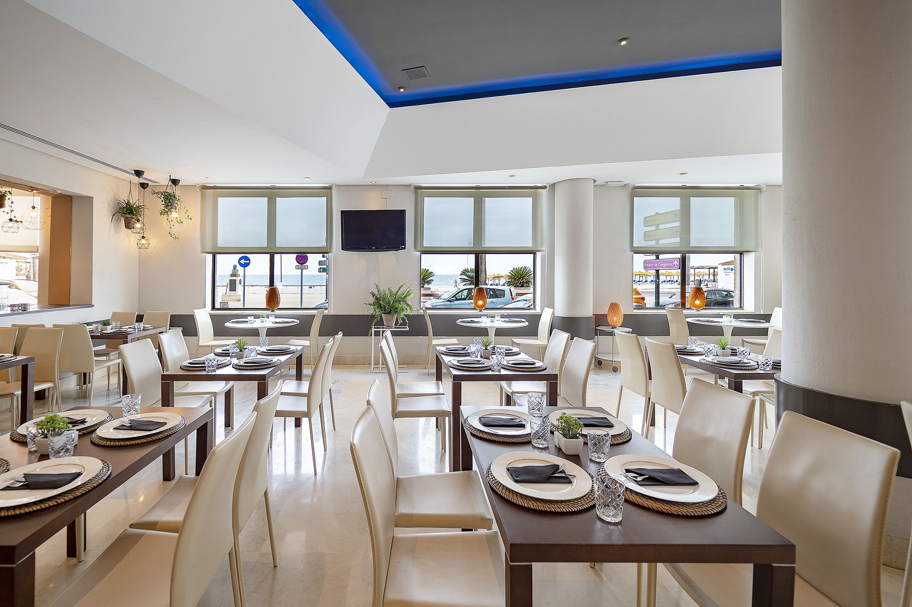 Un comedor moderno con mesas dispuestas para el servicio, amplias ventanas que ofrecen vistas al mar y un televisor de pantalla plana en la pared.