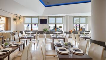 Un comedor moderno con mesas dispuestas para el servicio, amplias ventanas que ofrecen vistas al mar y un televisor de pantalla plana en la pared.