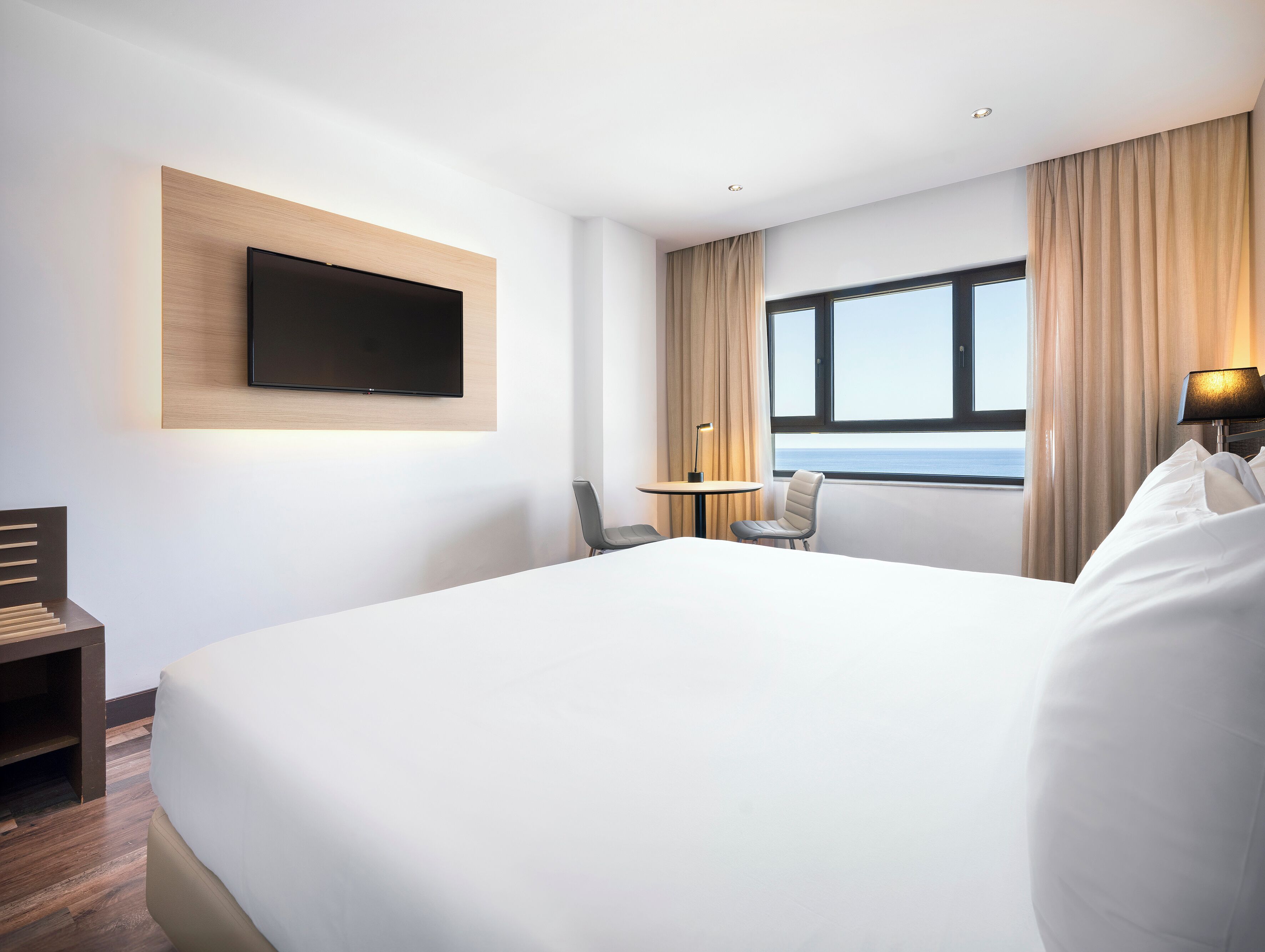 Una habitación de hotel moderna con una cama grande, un televisor de pantalla plana montado sobre un panel de madera, una mesita con una silla y vistas al mar a través de una ventana con cortinas.