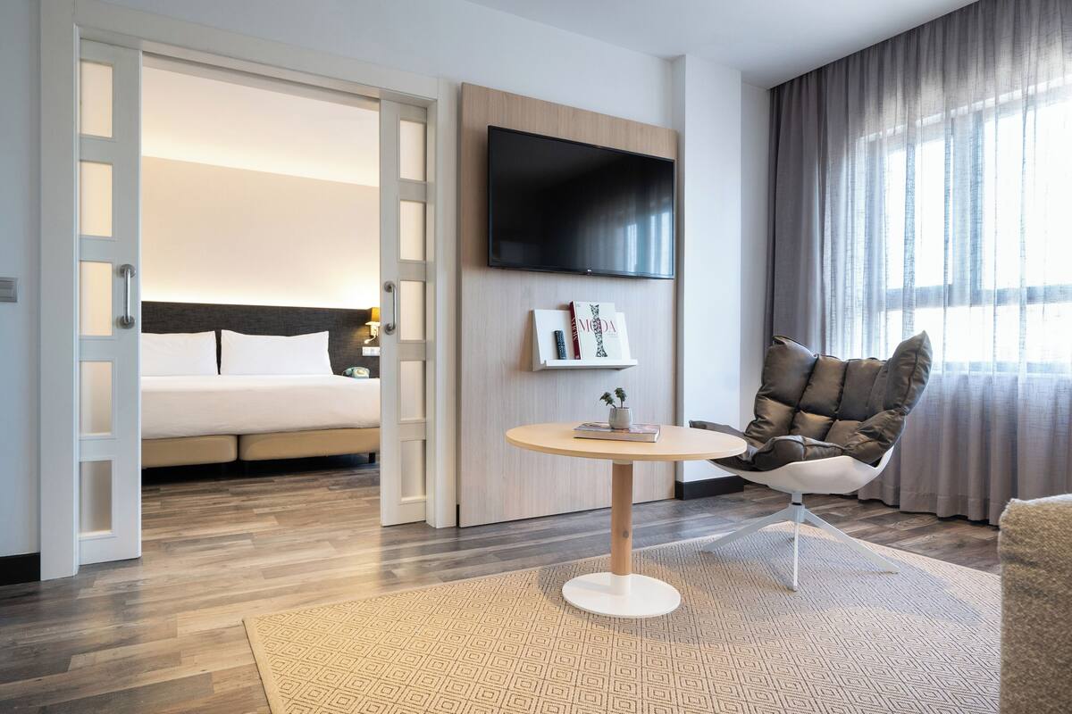 A modern hotel room with a bed, a TV mounted on the wall, a round table, and a comfortable chair.