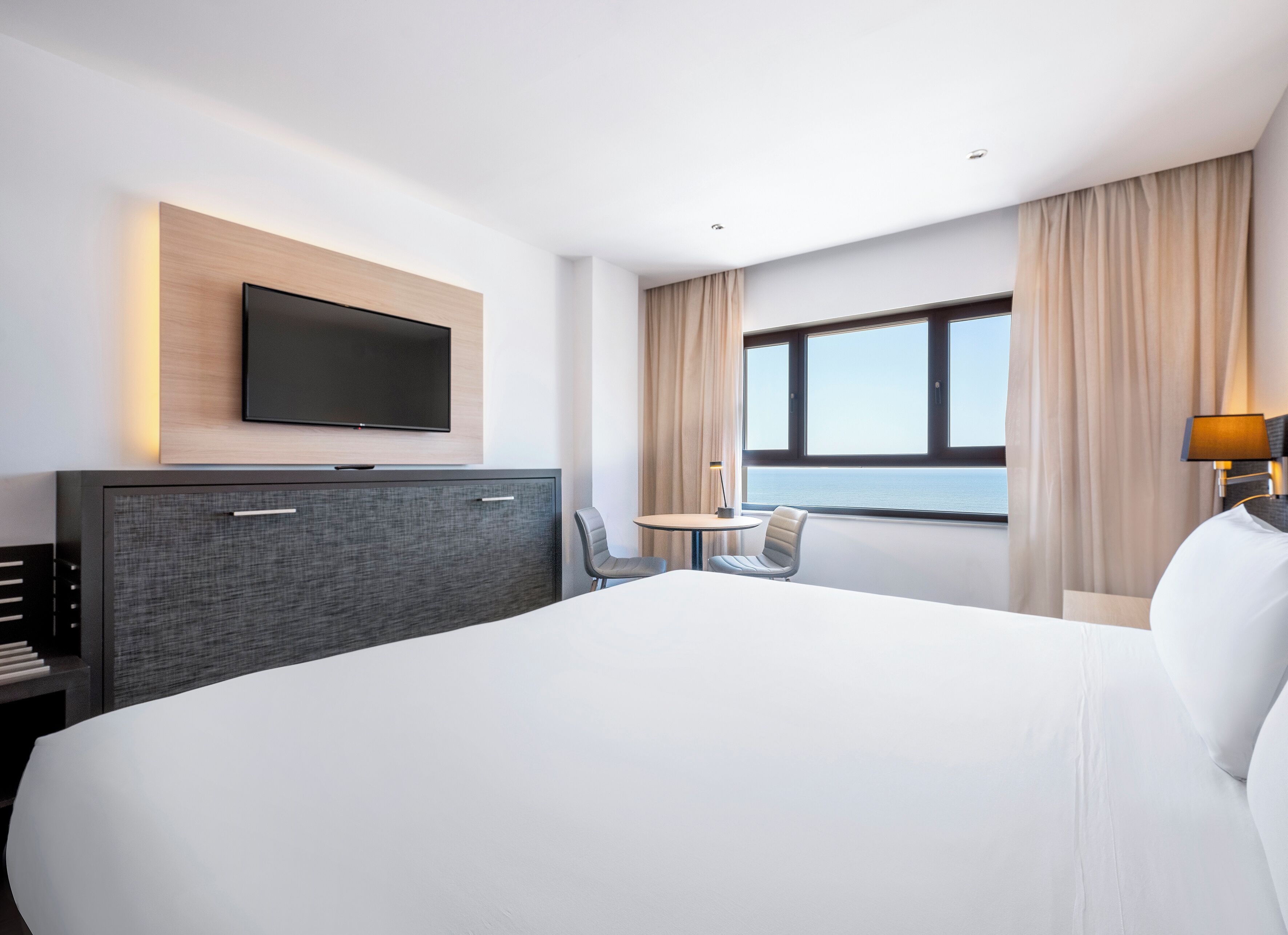 A modern hotel room with a large bed, a flat-screen TV mounted on the wall, a small table with two chairs, and a window with a view of the sea.