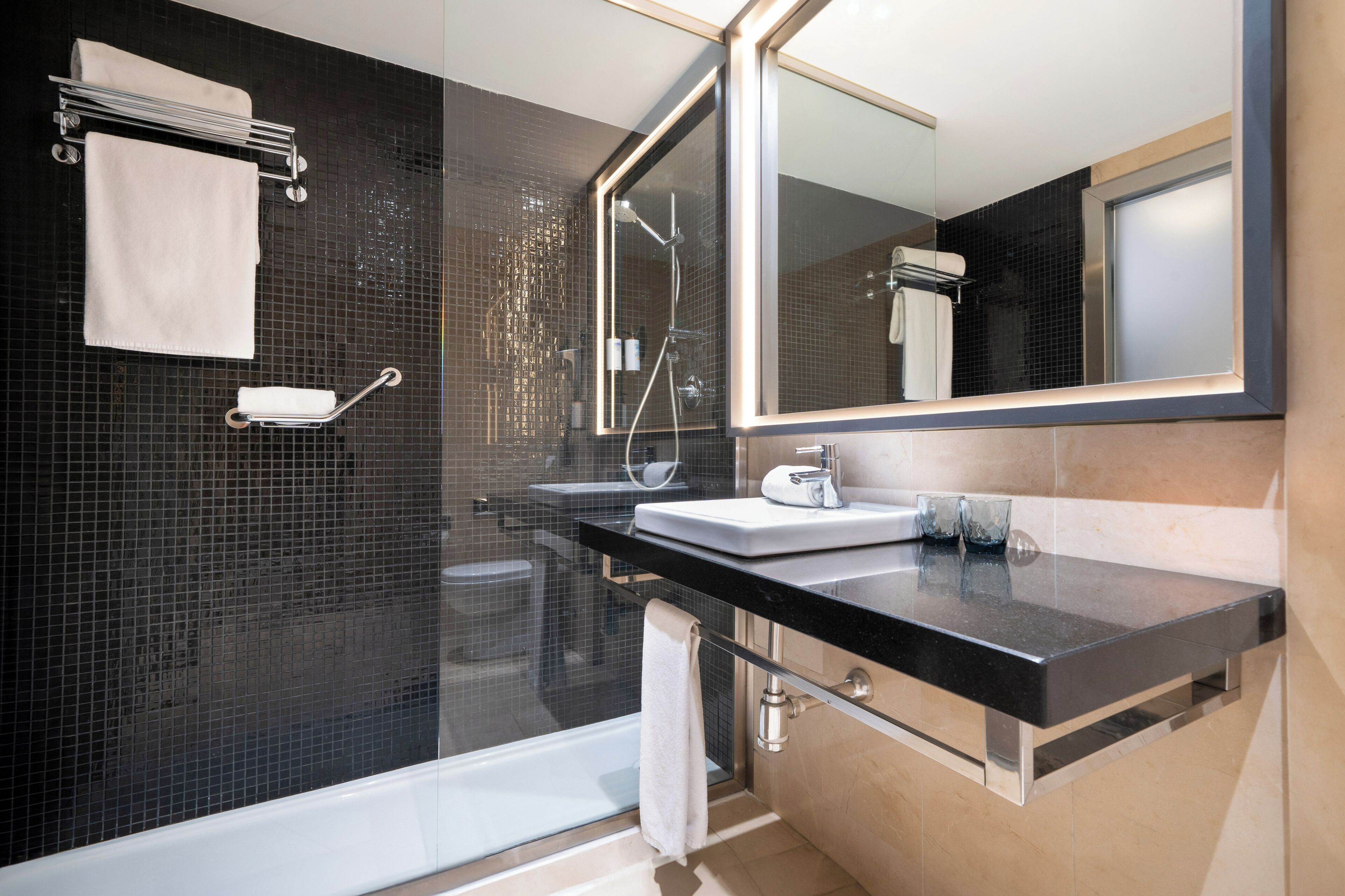 Un baño moderno con ducha de vidrio, lavamanos sobre encimera negra y un espejo grande.