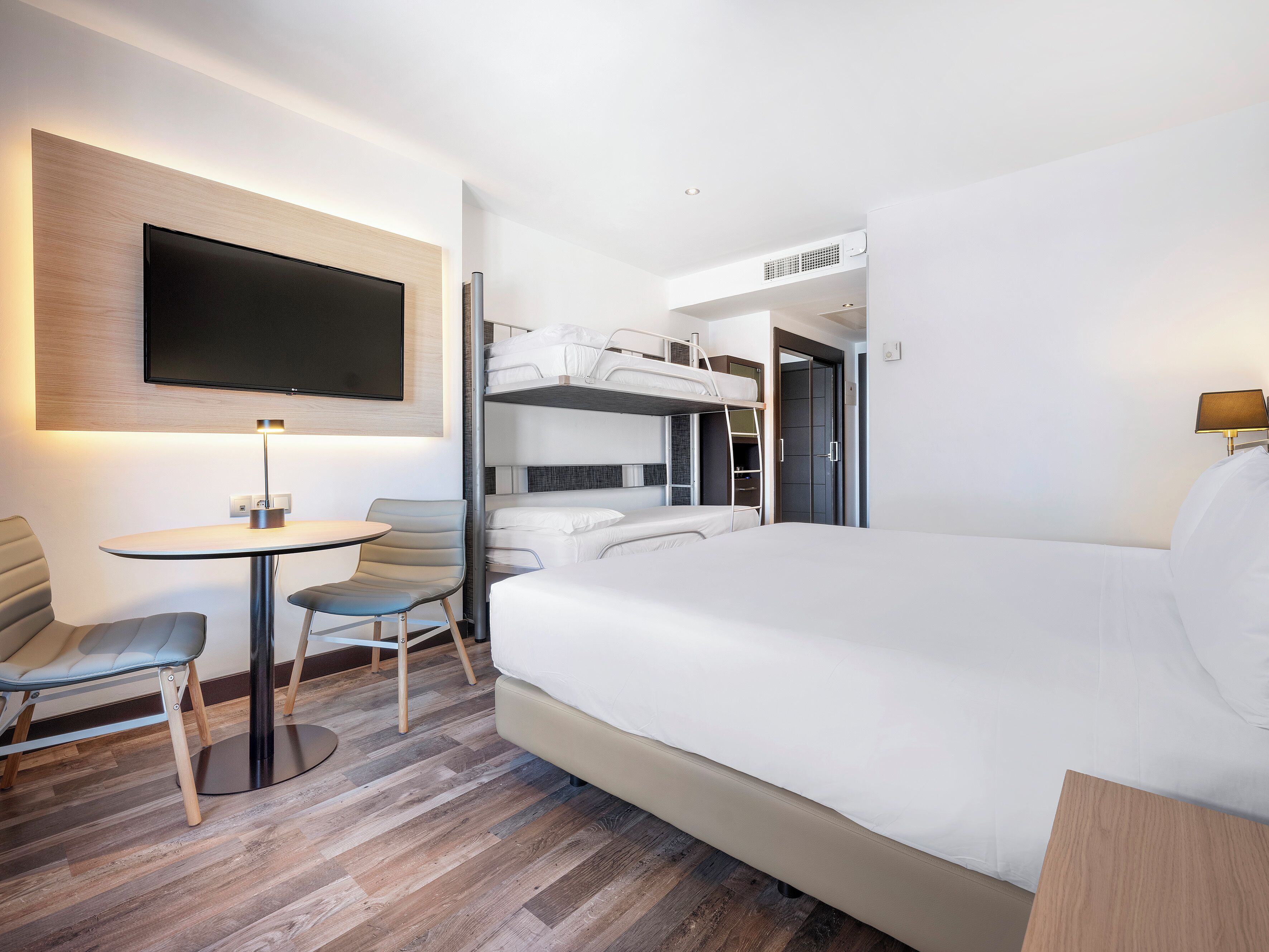 A modern hotel room with a large bed, a desk with a chair, a TV mounted on the wall, and a bunk bed in the background.