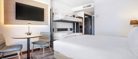 A modern hotel room with a large bed, a desk with a chair, a TV mounted on the wall, and a bunk bed in the background.
