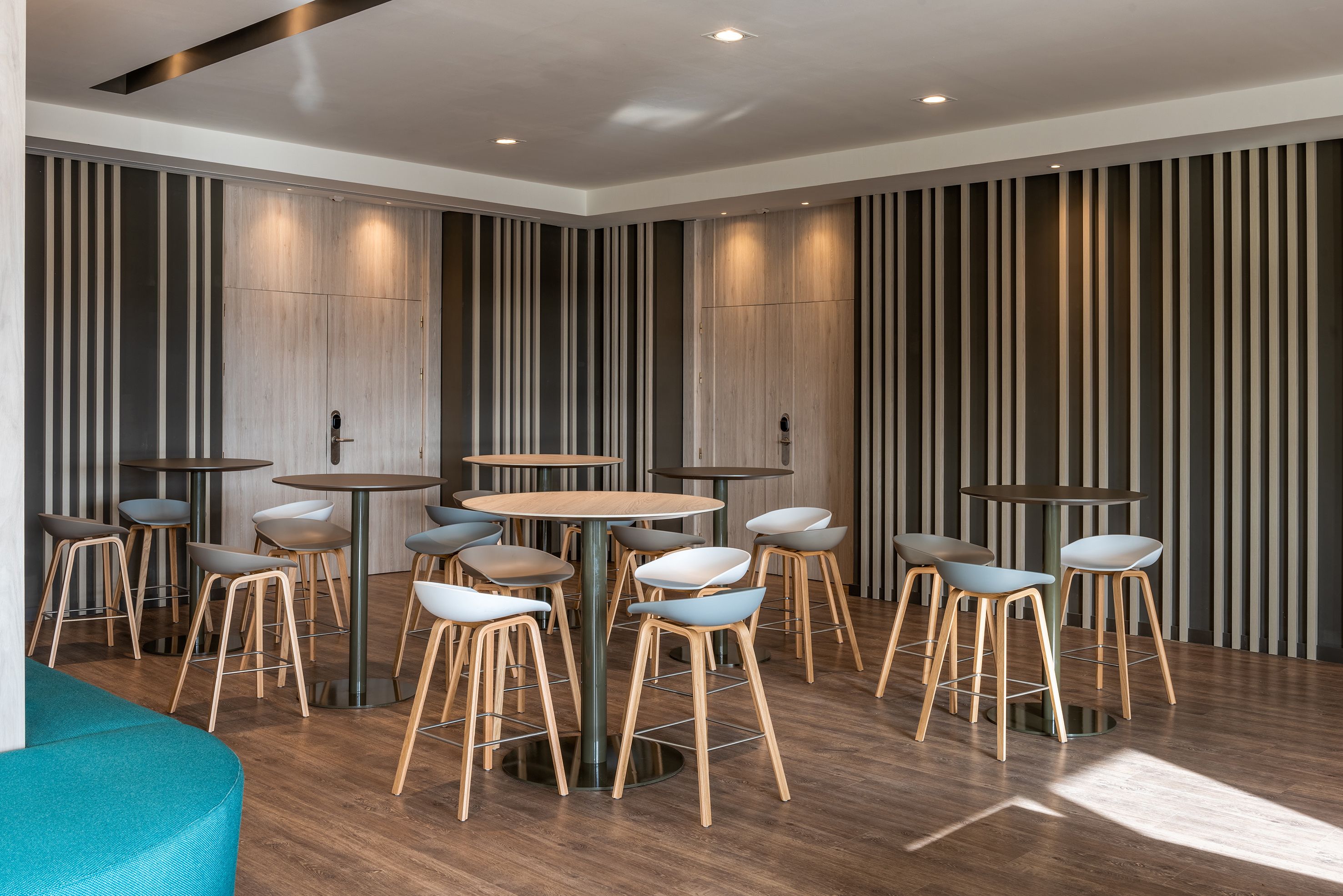 A modern interior with round tables and wooden stools, vertical wooden slats on the walls, and a teal-colored seating area.