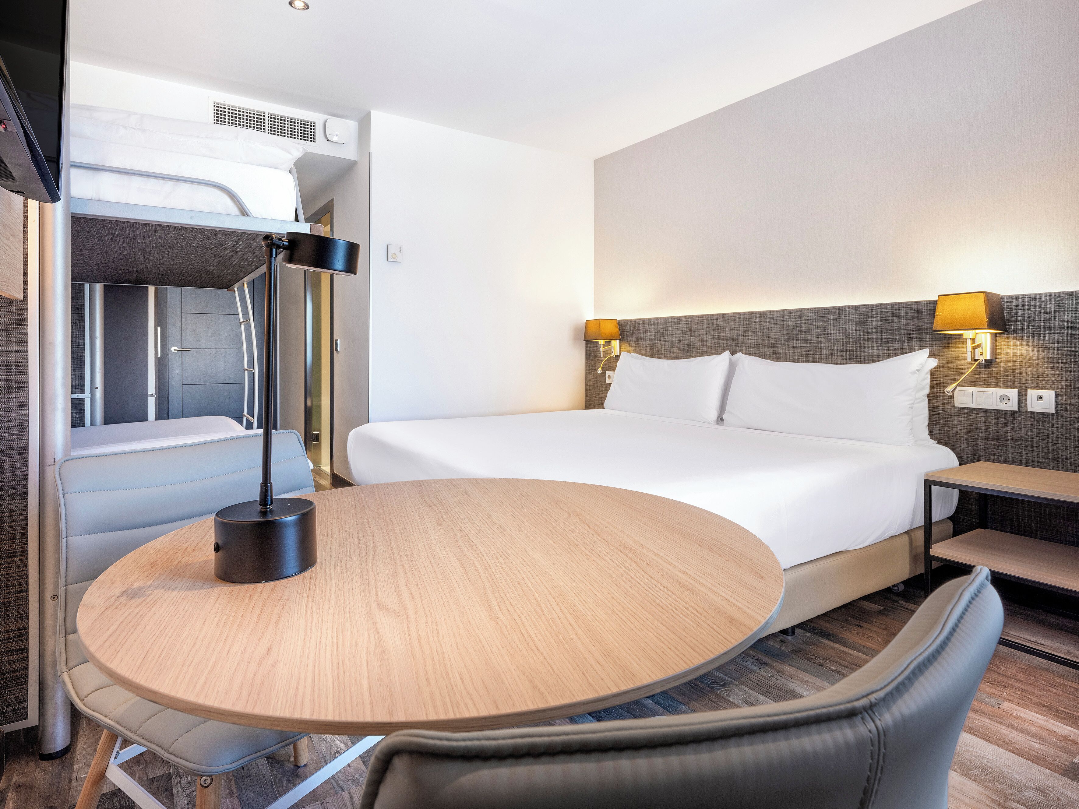 Una habitación de hotel moderna con una cama, mesita de noche, lámpara y una pequeña zona de estar.
