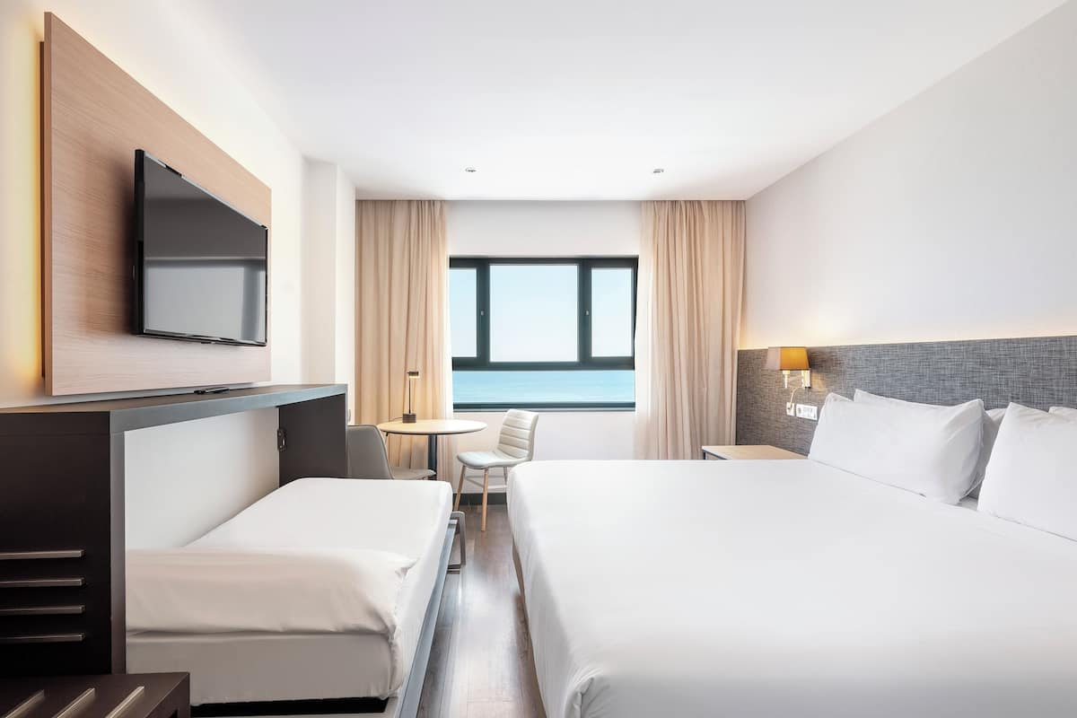 A hotel room with two beds, a TV, a desk with a chair, a lamp, and a view of the sea.