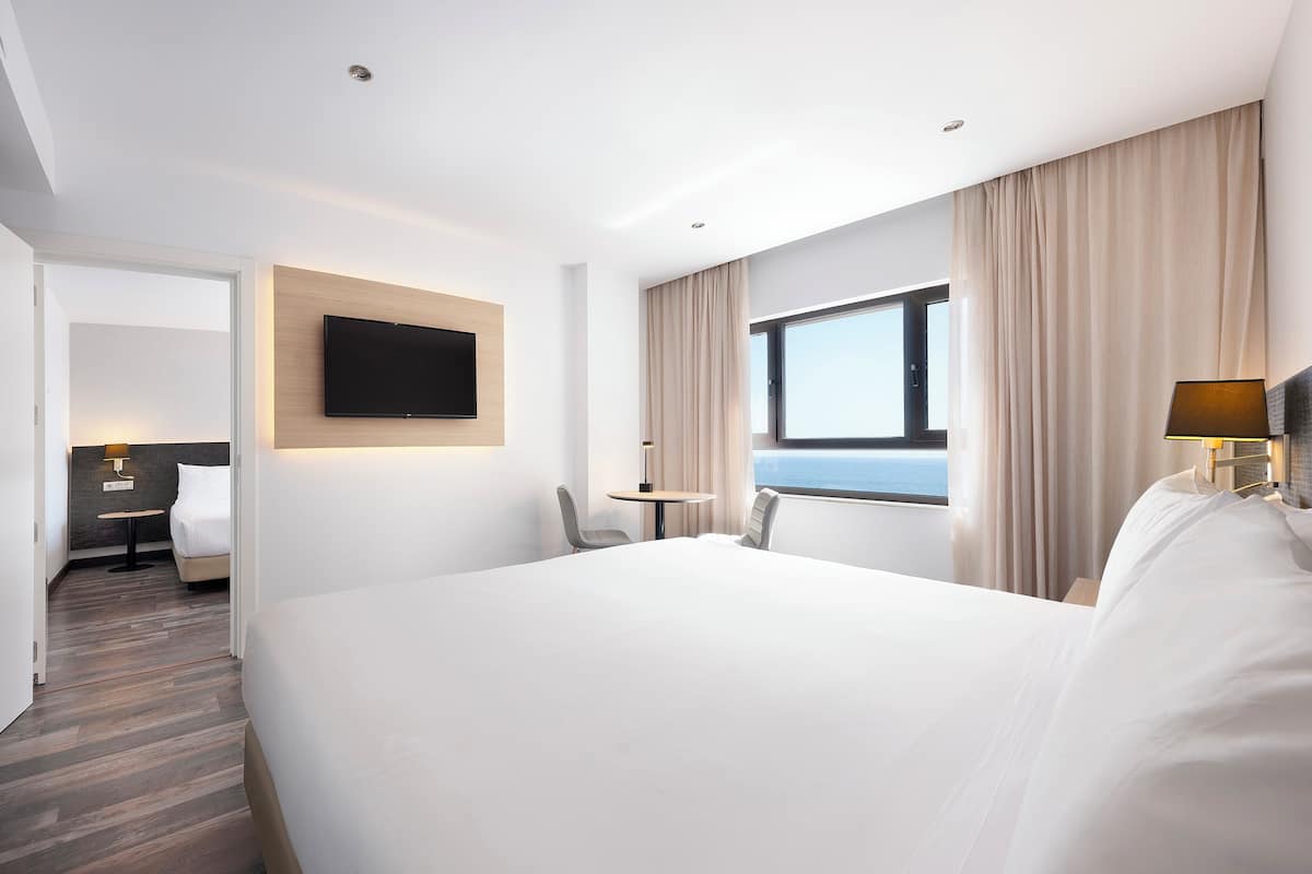 A hotel room with a large bed, a TV mounted on the wall, a small table, and a view of the sea through a window.
