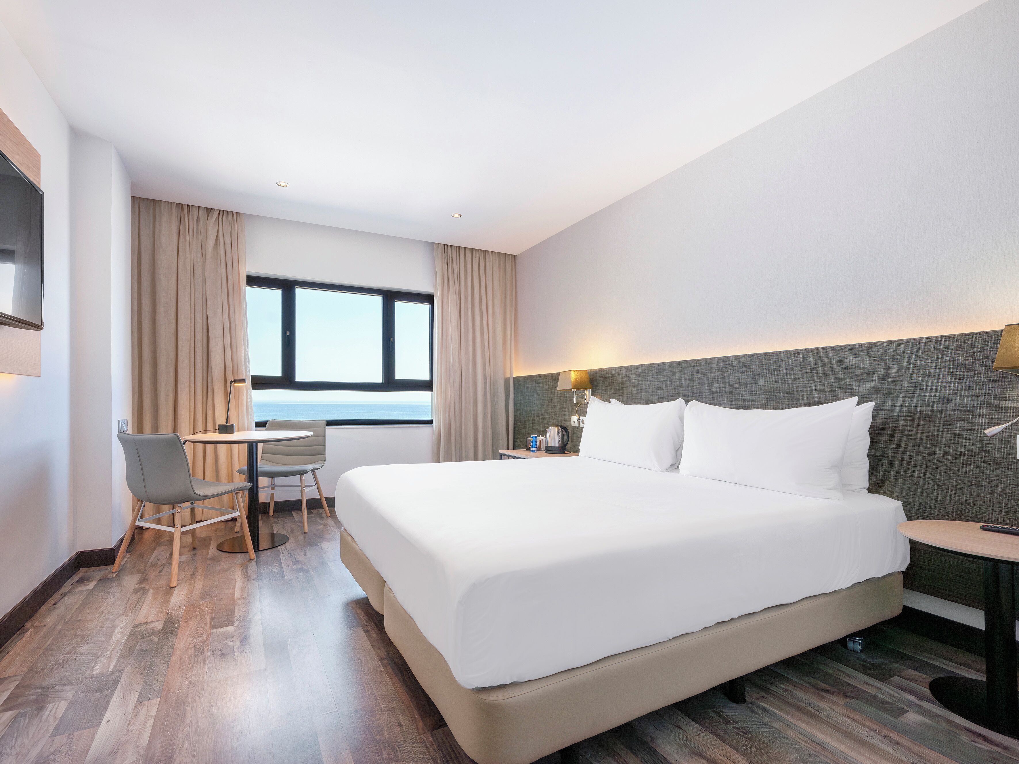 Une chambre d’hôtel avec un grand lit, un bureau, une chaise, une fenêtre avec des rideaux et une lampe fixée au mur.