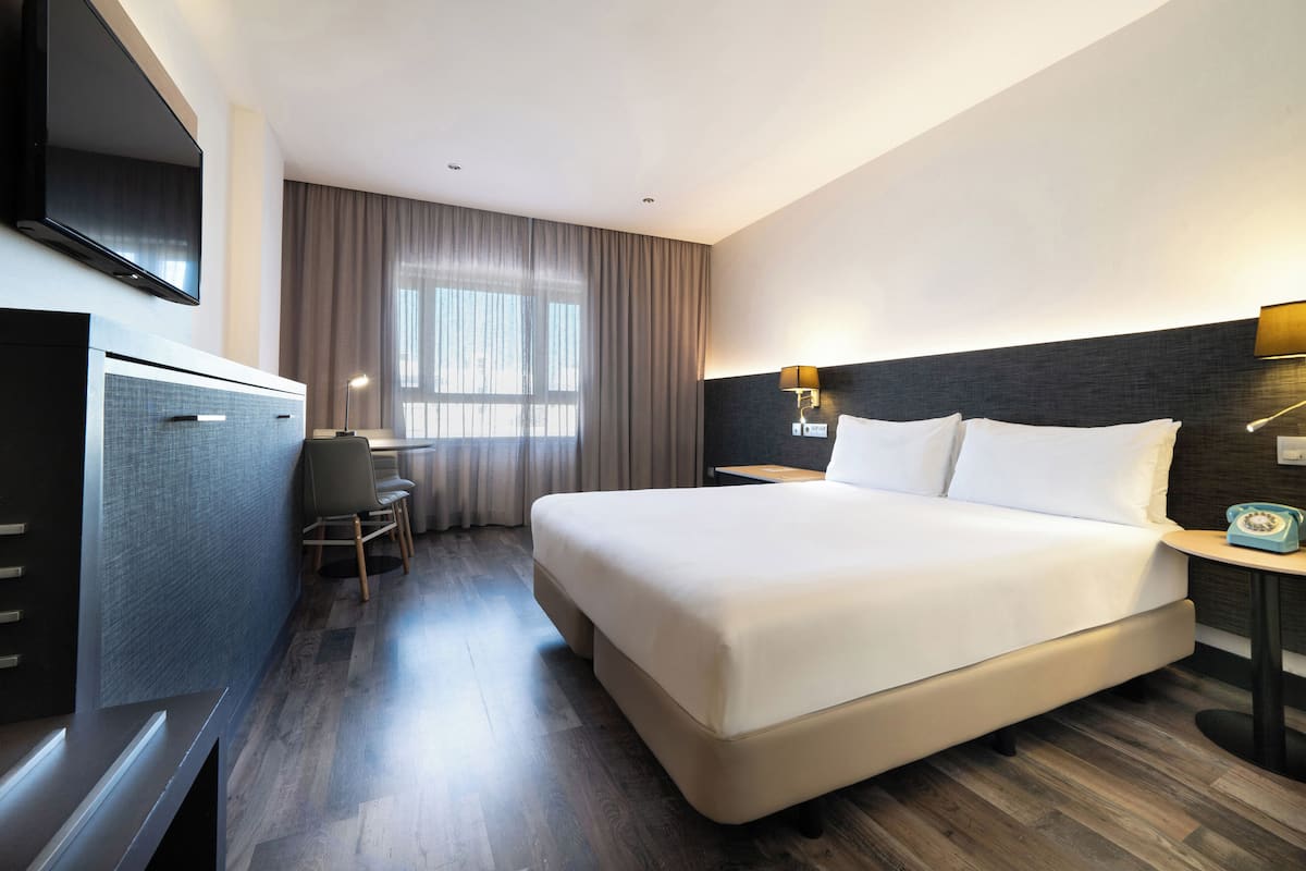 A hotel room with a large bed, a desk with a chair, a television mounted on the wall, and a window with curtains.